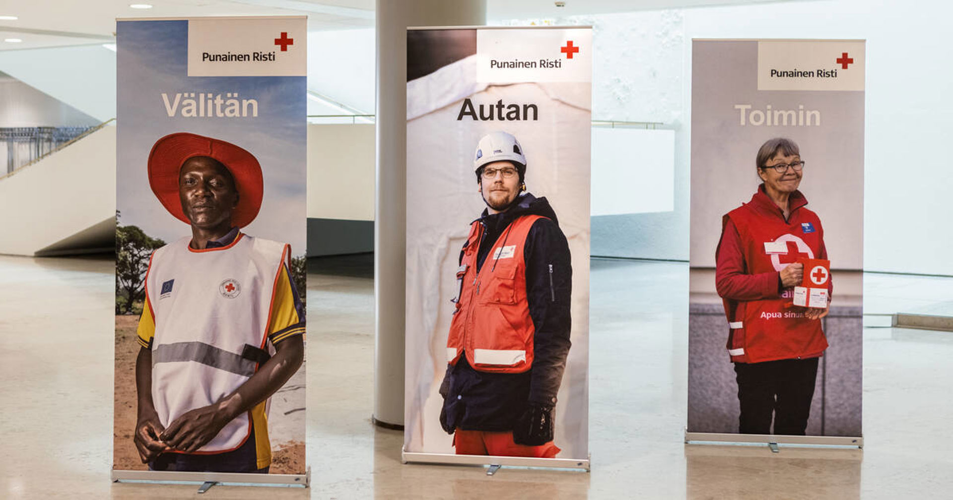 Aulassa pystyssä kolme roll up -esitettä, joissa on punaisen ristin vapaaehtoinen ja teksti välitän, autan, toimin.