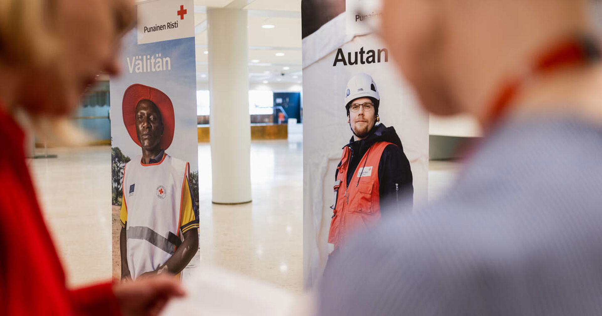 Punaisen Ristin rollupit, joissa lukee autan ja välitän, aulassa. Kaksi henkilöä lukee esitettä etualalla.