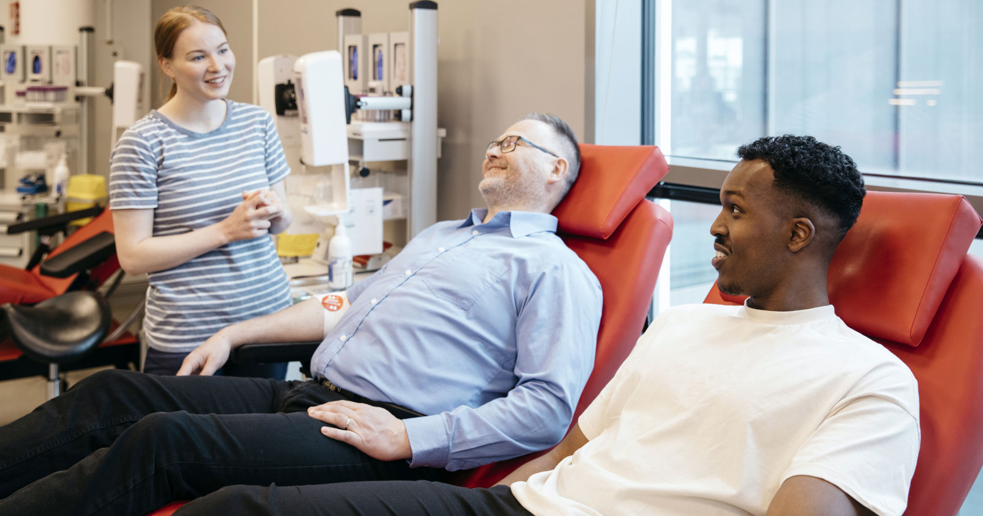 Kaksi henkilöä hymyilee hoitajalle verenluovutuksen jälkeen. Heidän käsivarsissaan on siteet ja veripalvelun tarra.