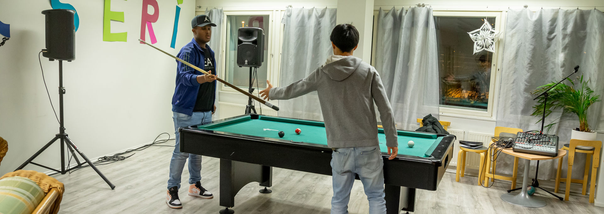 Nuori henkilö heittää toiselle nuorelle henkilölle biljardikepin. He seisovat biljardipöydän ääressä.