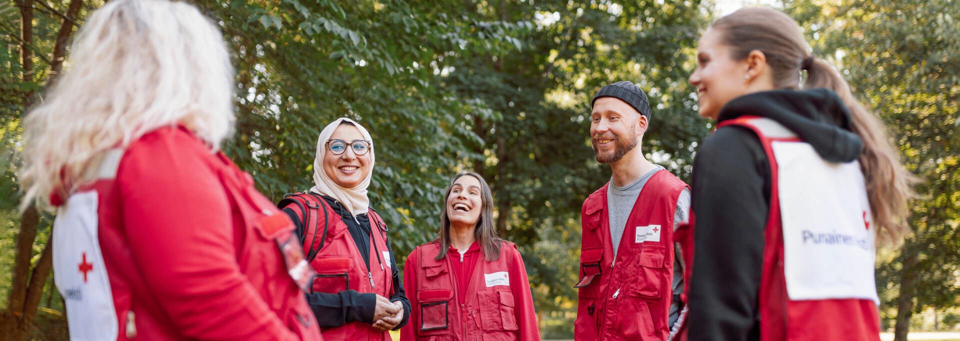 Viisi Punaisen Ristin vapaaehtoista seisovat ringissä ja juttelevat iloisesti keskenään. He ovat ulkona ja heillä on päällään punaiset vapaaehtoisliivit.