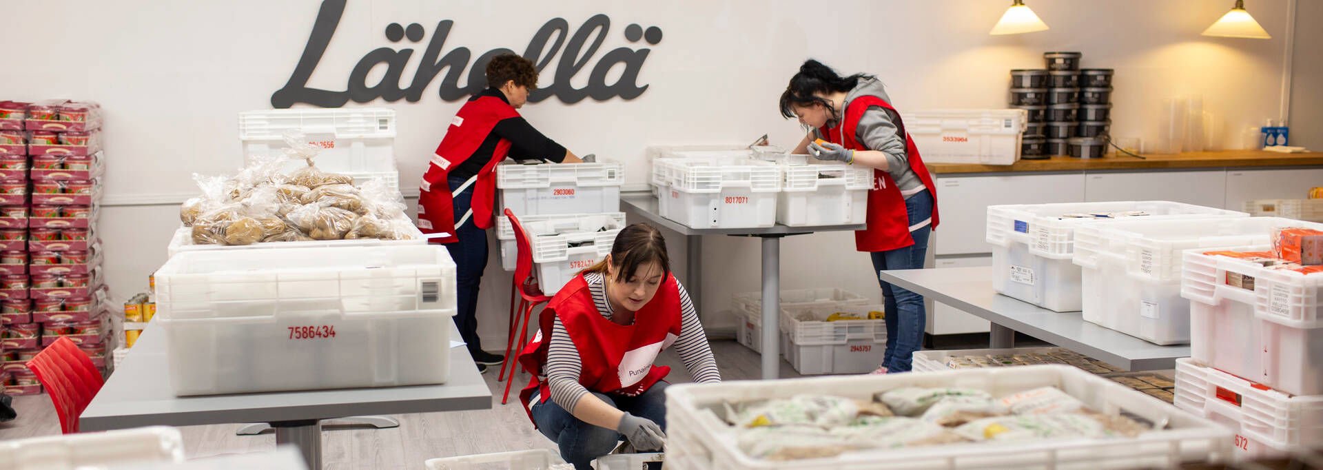 Kolme Punaisen Ristin vapaaehtoista lajittelevat ruoka-avussa jaettavaa ruokaa. Heillä on päällään punaiset vapaaehtoisliivit.