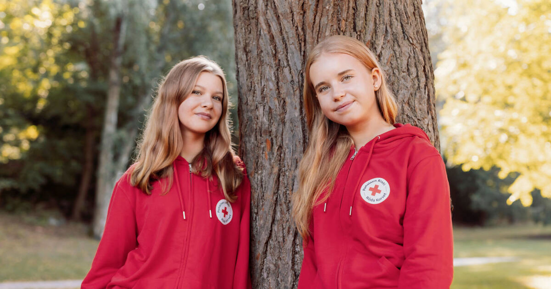 Kaksi nuorta nojailee puuhun ulkona. Heillä on päällään punaiset Punaisen Ristin hupparit.