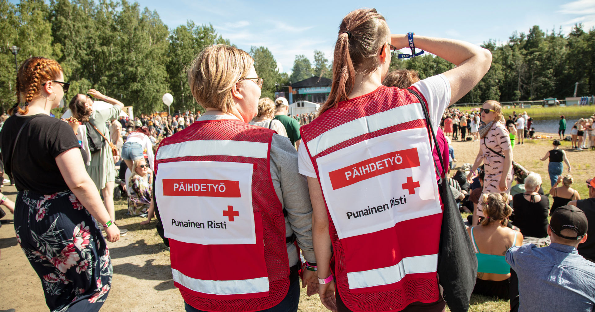 Kaksi Punaisen Ristin vapaaehtoista seisoo selin kameraan festareilla. Heillä on päällään vapaaehtoisliivit, joissa lukee Päihdetyö.