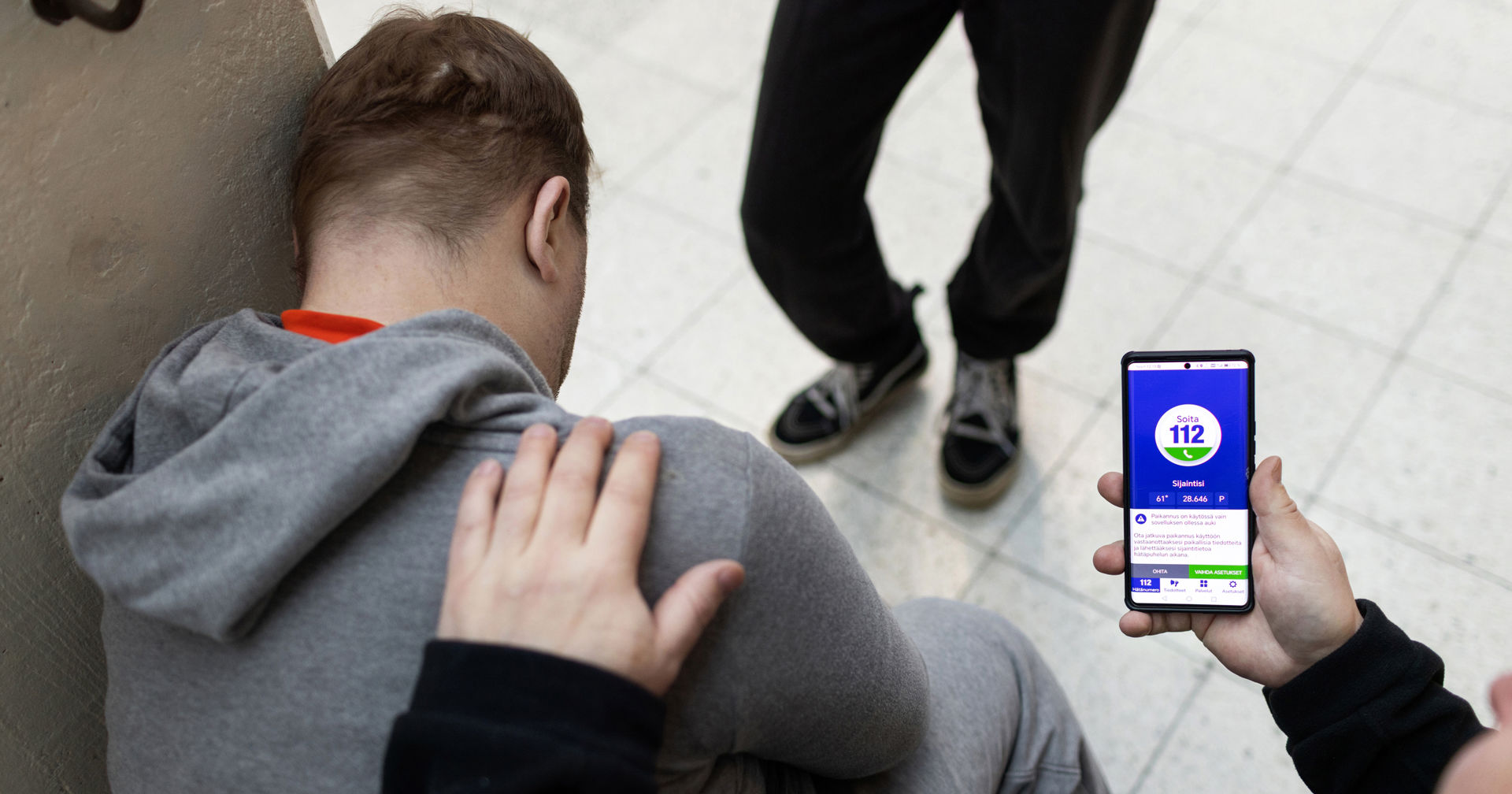 Henkilö nojaa seinään. Toinen pitää kädellä häntä olasta ja on avannut puhelimestaan 112 aplikaation.