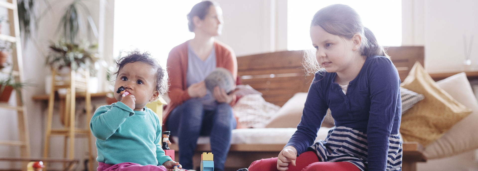 Kaksi lasta leikkii leluilla etualalla, ja pieni lapsi on laittanut lelun suuhunsa. Taustalla vanhempi lukee lehtä..
