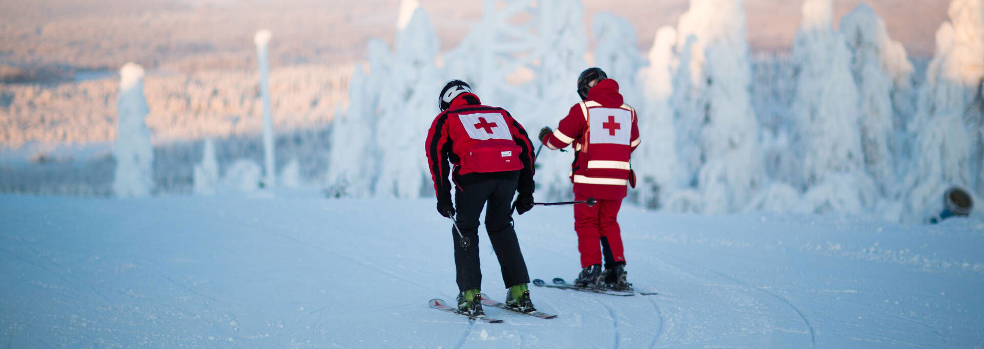 Laskettelurinteessä kaksi punapukaista laskettelijaa Punaisen Ristin liivit päällään.