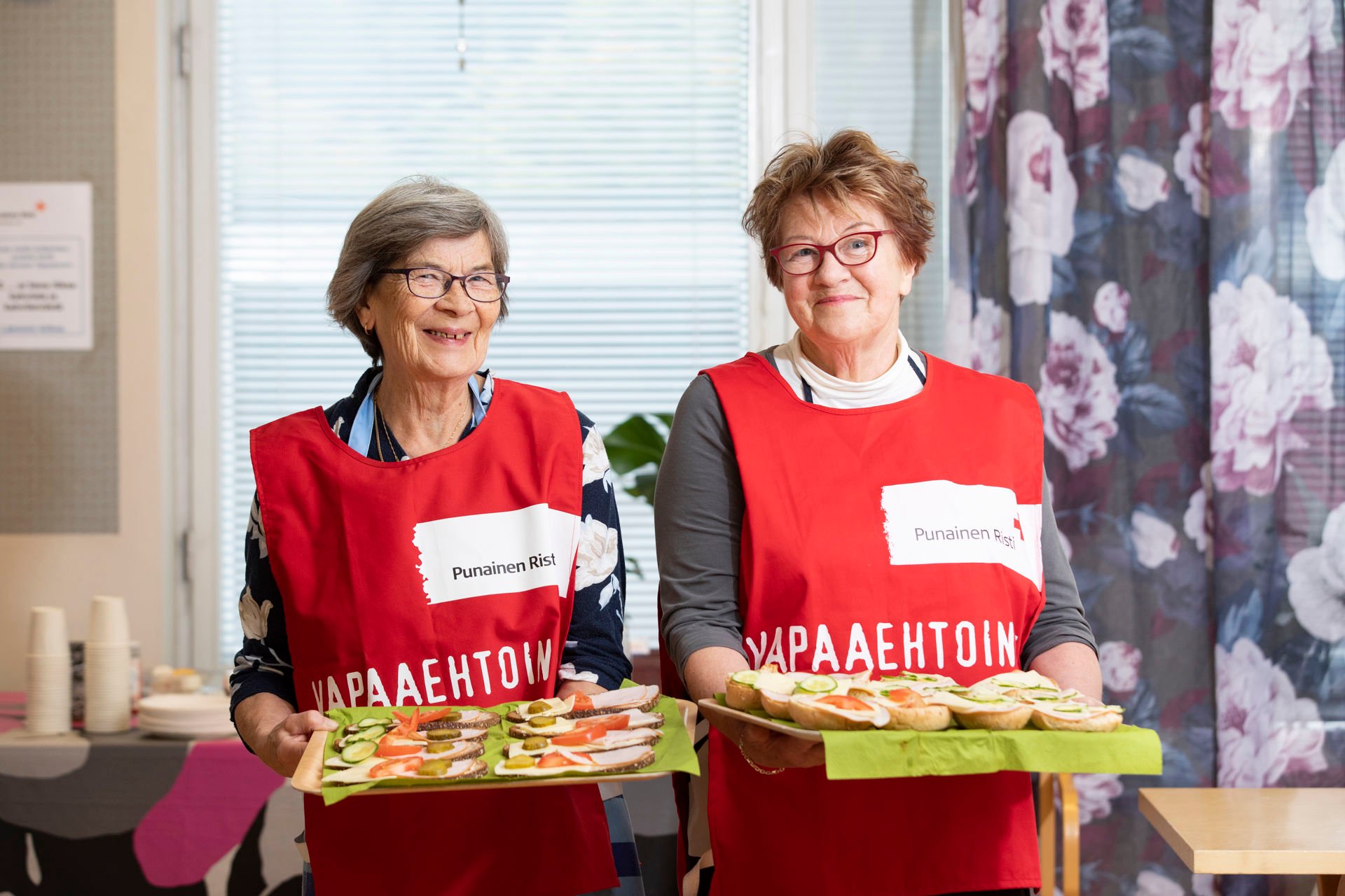 Kaksi vapaaehtoista hymyilee kameralle ja pitää käsissään tarjottimia, joilla on voileipiä.