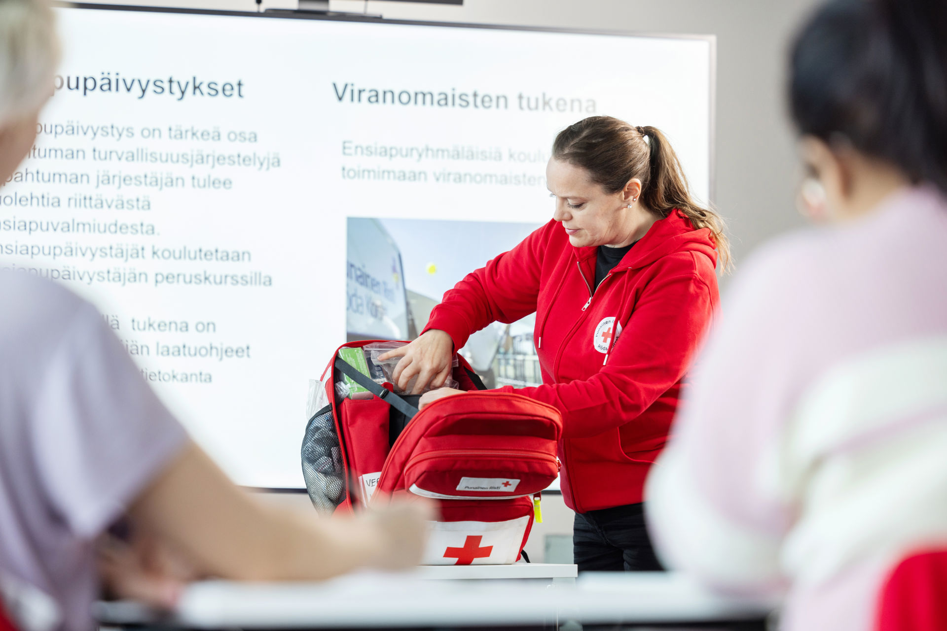 Suomen Punaisen Ristin kouluttaja etsii jotain ensiapurepusta valkokankaan edessä.