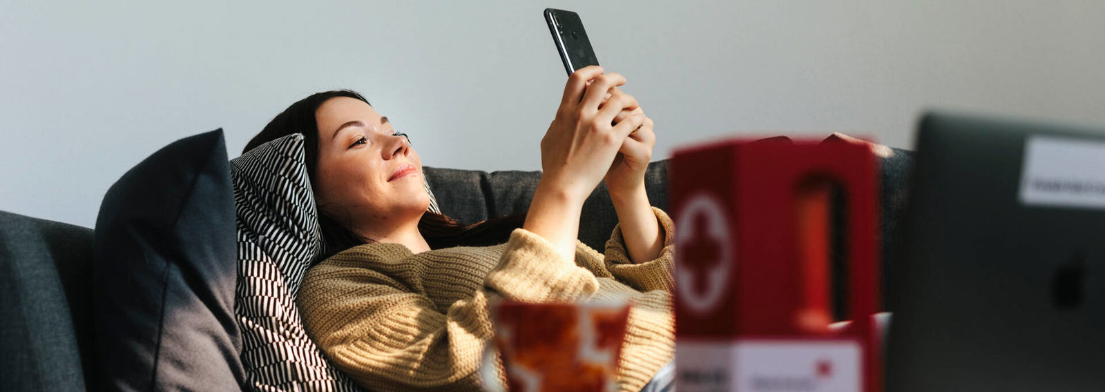 Henkilö makaa sohvalla puhelin kädessään. Etualalla on Punaisen Ristin logo keräyslippaassa.