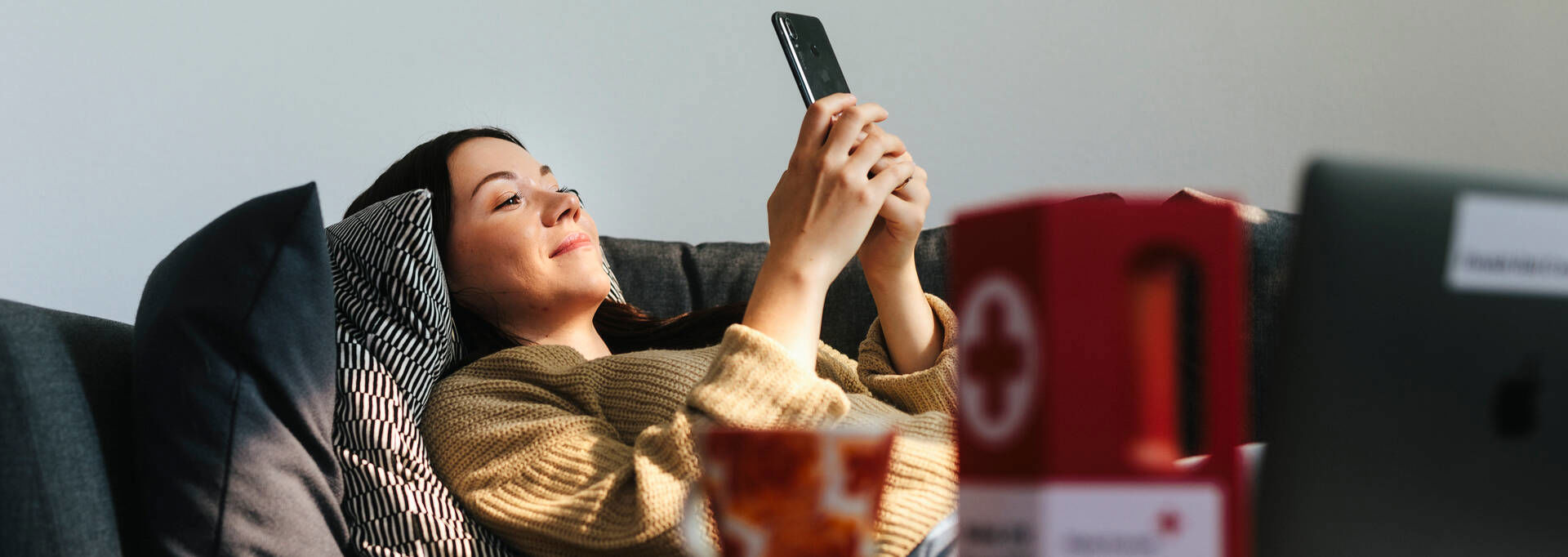 Henkilö makaa sohvalla puhelin kädessään. Etualalla on Punaisen Ristin logo keräyslippaassa.