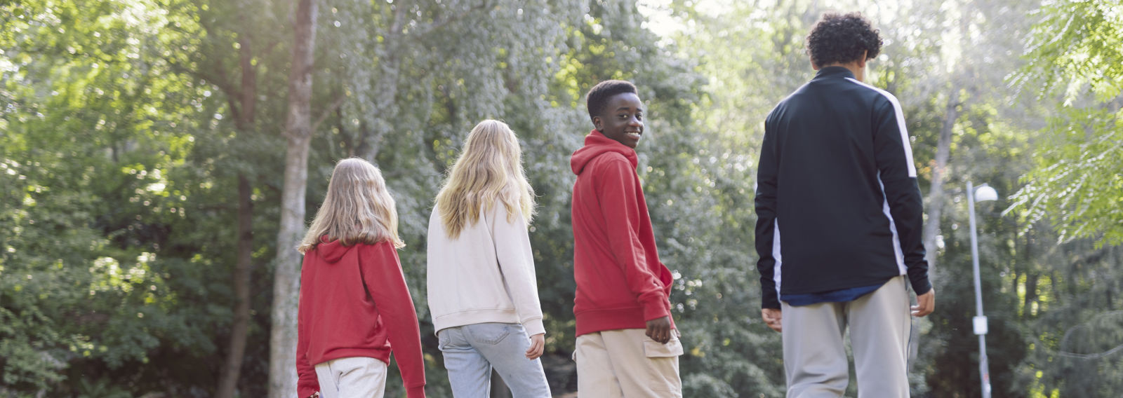 Neljä nuorta kulkee puistossa selät kohti kameraa. Yksi heistä on kääntänyt päänsä ja hymyilee kameraan.