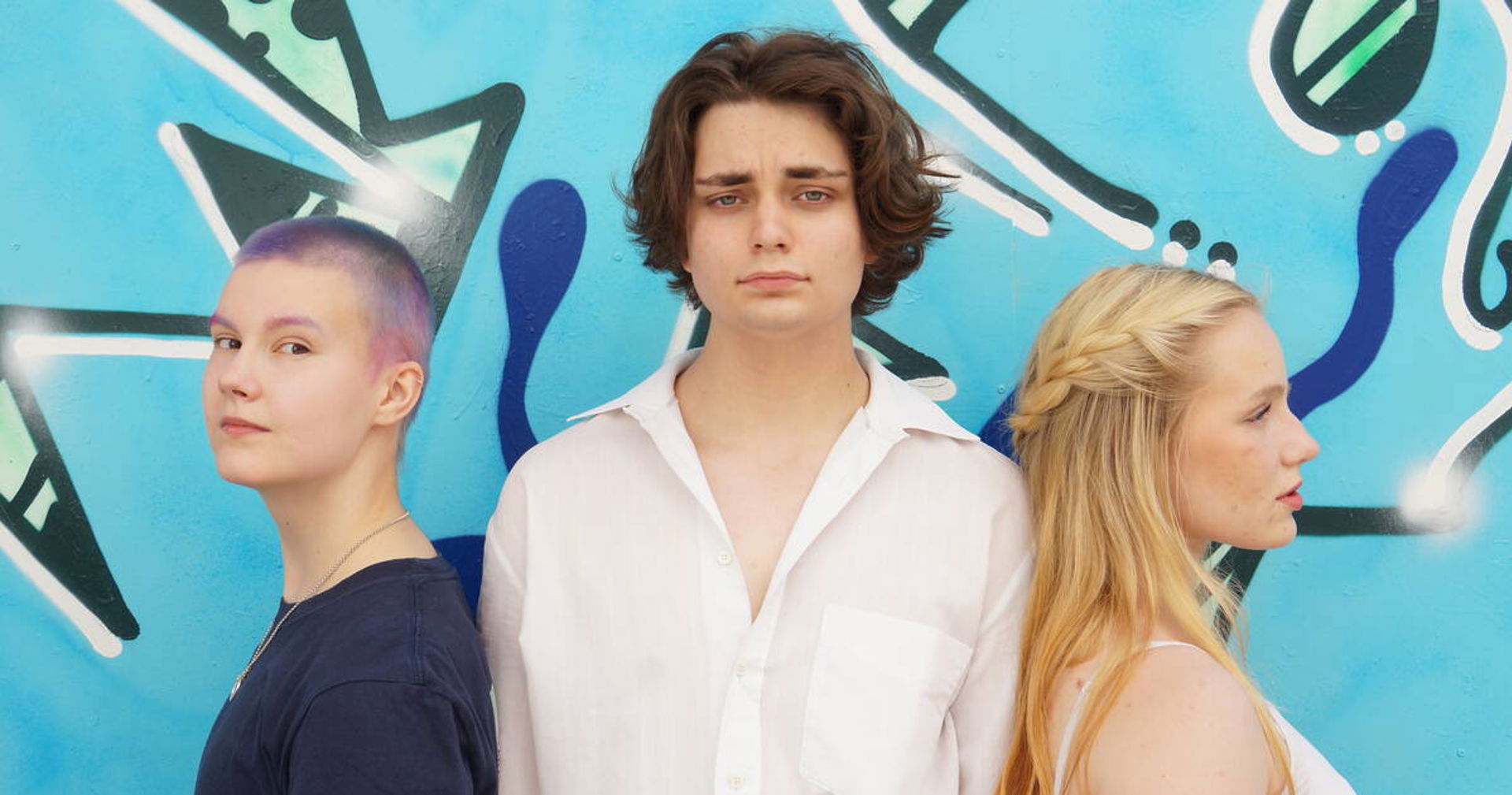 Kuvassa on kolme nuorta selät vastakkain. Kaksi nuorista katsoo kameraan ja yksi sivulle.