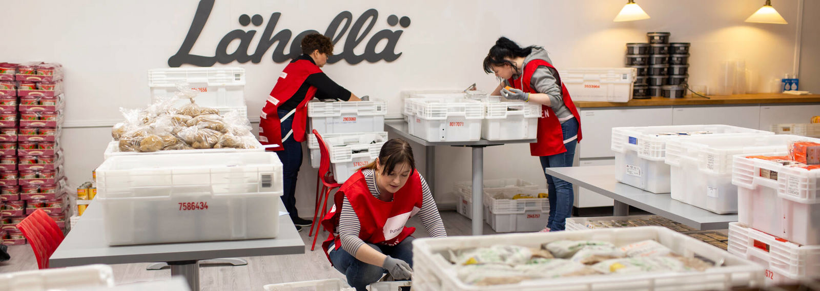 Kolme Punaisen Ristin vapaaehtoista lajittelevat ruoka-avussa jaettavaa ruokaa. Heillä on päällään punaiset vapaaehtoisliivit.