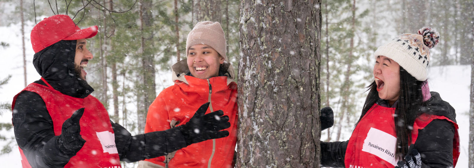 Kolme nuorta ovat puun ympärillä ja nauravat yhdessä. Kahdella heistä on päällään Punaisen Ristin vapaaehtoisliivit.