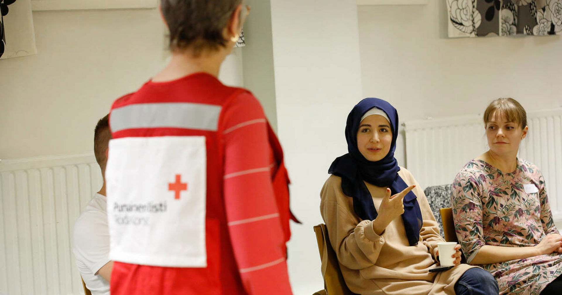 Kuvassa selin kameraan on vapaaehtoiskouluttaja, jolla on päällään punainen vapaaehtoisliivi. Lisäksi kuvassa istuu kaksi nuorta vapaaehtoista kasvot kameraan päin.