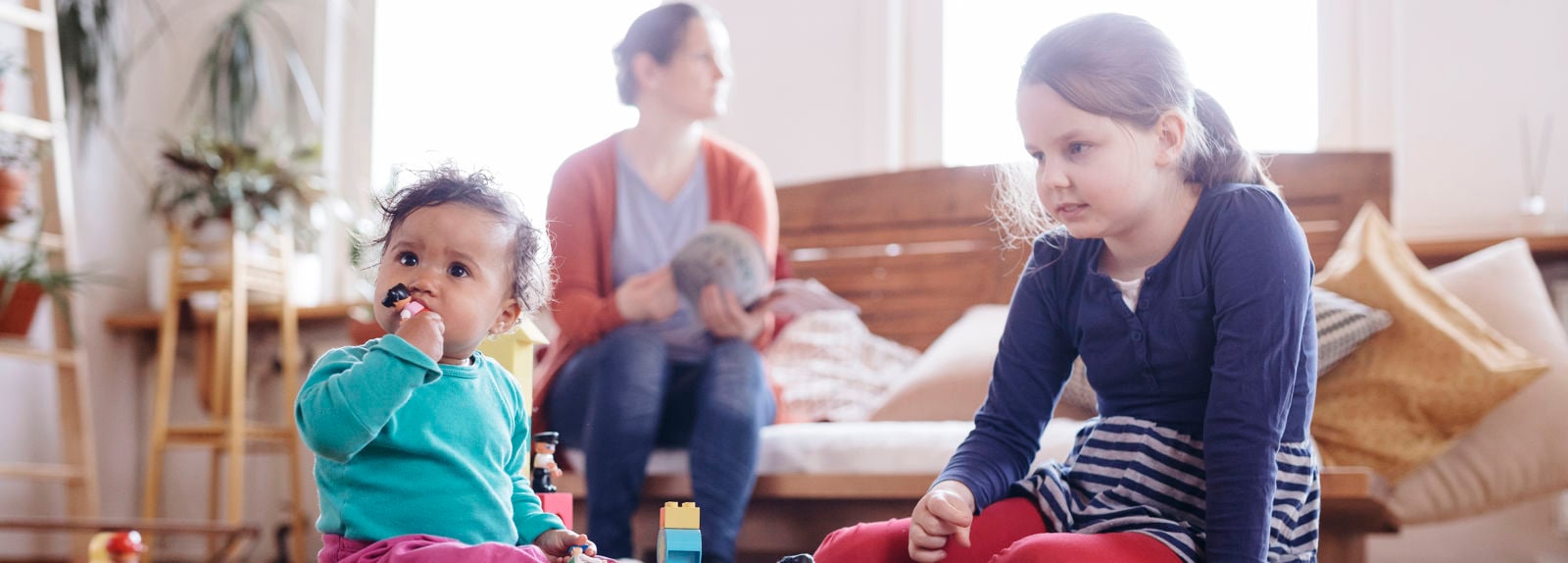 Kaksi lasta leikkii leluilla etualalla, ja pieni lapsi on laittanut lelun suuhunsa. Taustalla vanhempi lukee lehtä..