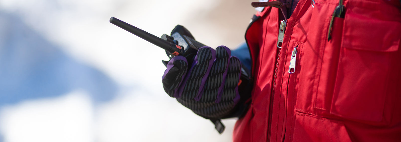 Punaiseen liiviin pukeutunut henkilö pitää radiopuhelinta hanskaisessa kädessään. Tausta on valkoinen ja luminen.