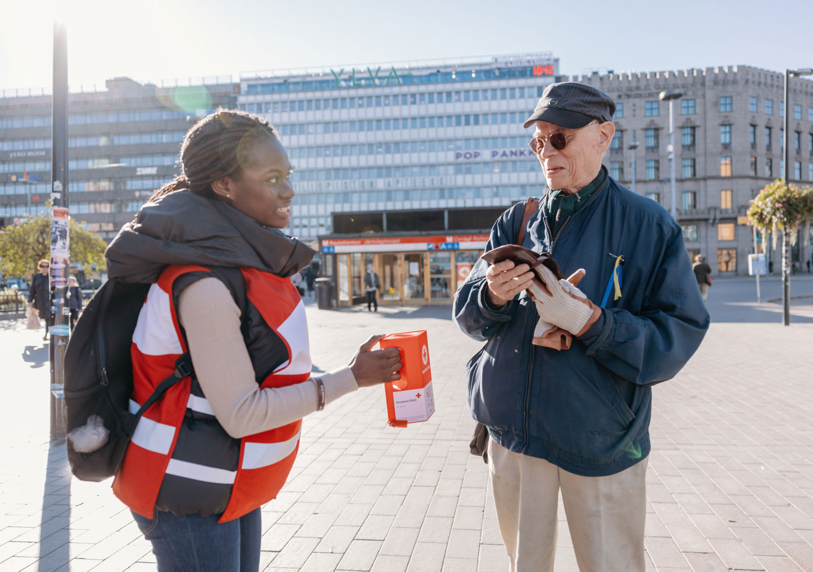 Hymyilevä henkilö laittaa rahaa Punaisen Ristin vapaaehtoisen keräyslippaaseen.
