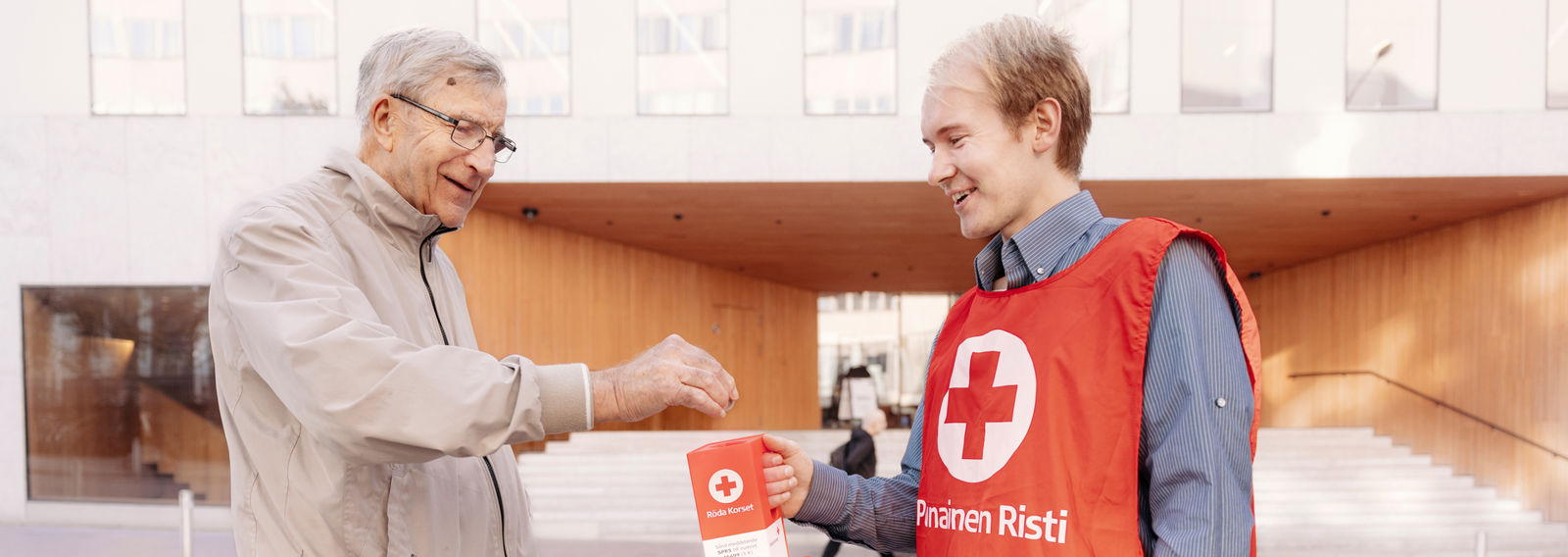 Hymyilevä henkilö laittaa rahaa Punaisen Ristin vapaaehtoisen keräyslokeroon.