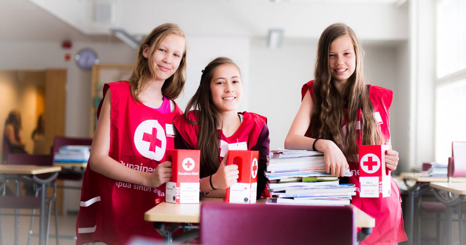 Kolme koululaista nojaa pulpetteihin keräysliivit päällään ja keräyslippaat käsissään.