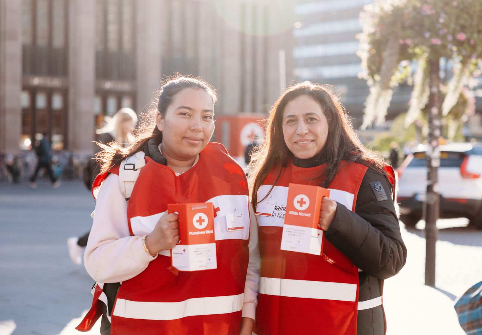 Kaksi lipaskerääjää hymyilevät vierekkäin keräyslippaat kädessään. 
