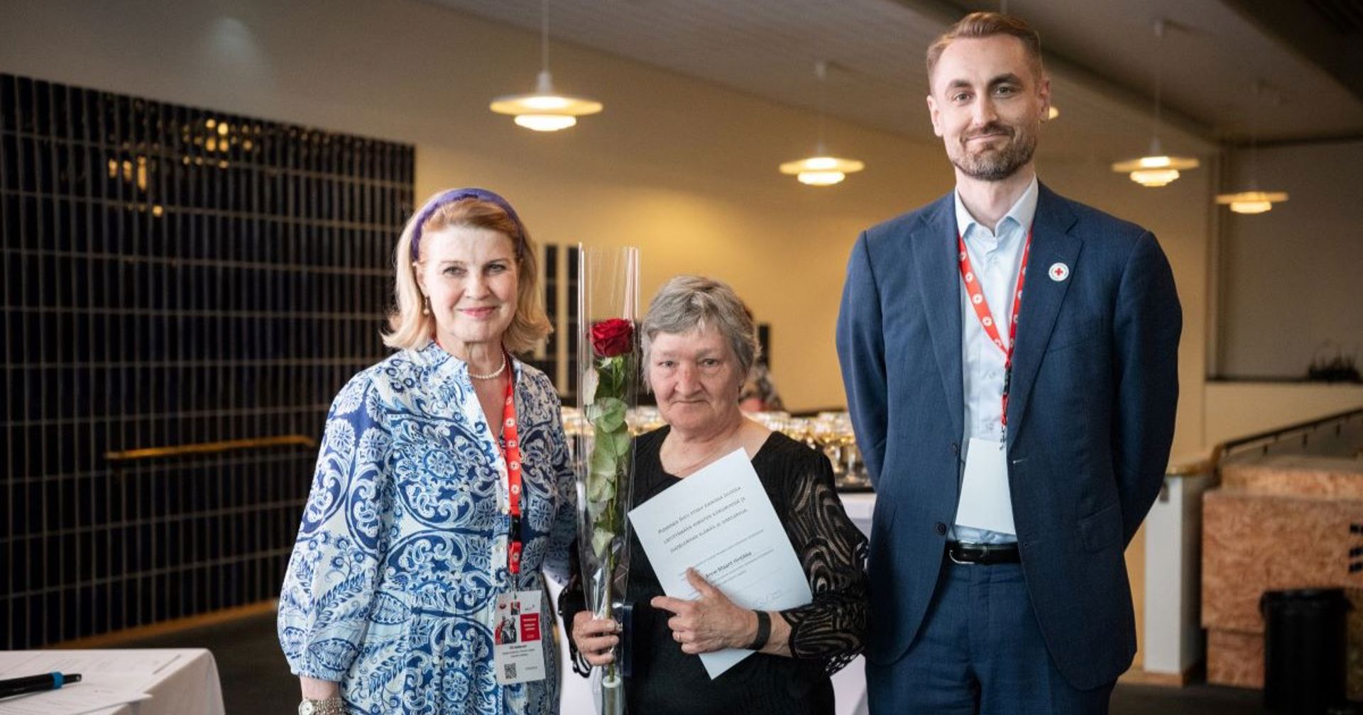 Kolme henkilöä palkitsemistilaisuudessa, keskellä ansiomitalin saanut henkilö pitää kukkaa ja kunniakirjaa.