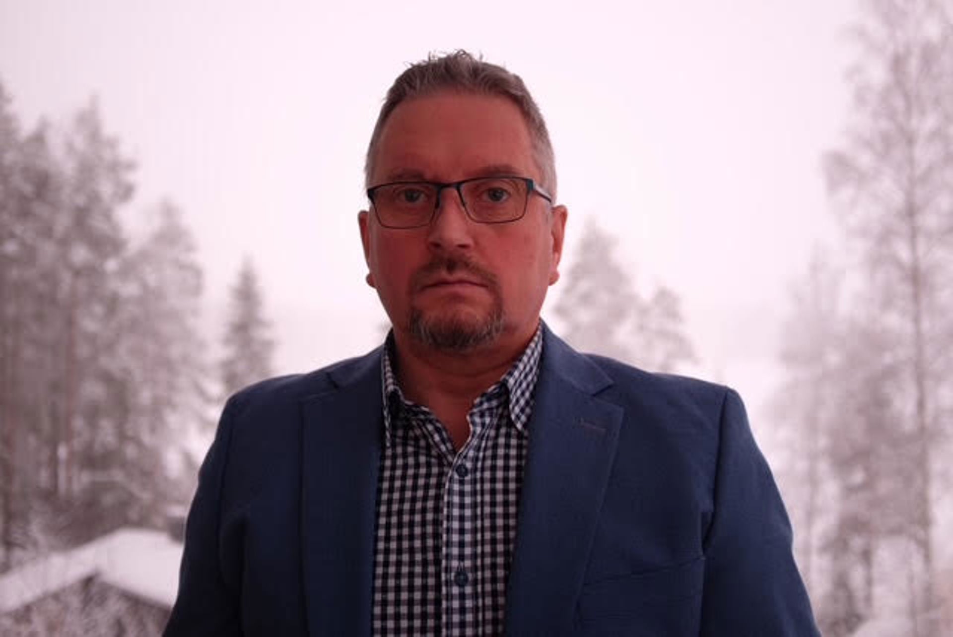 Heikki Väätämöinen katsoo kohti kameraa, päällään sininen bleiseri ja ruudullinen kauluspaita. Taustalla on talvimaisema, jossa näkyy lumisia puita.