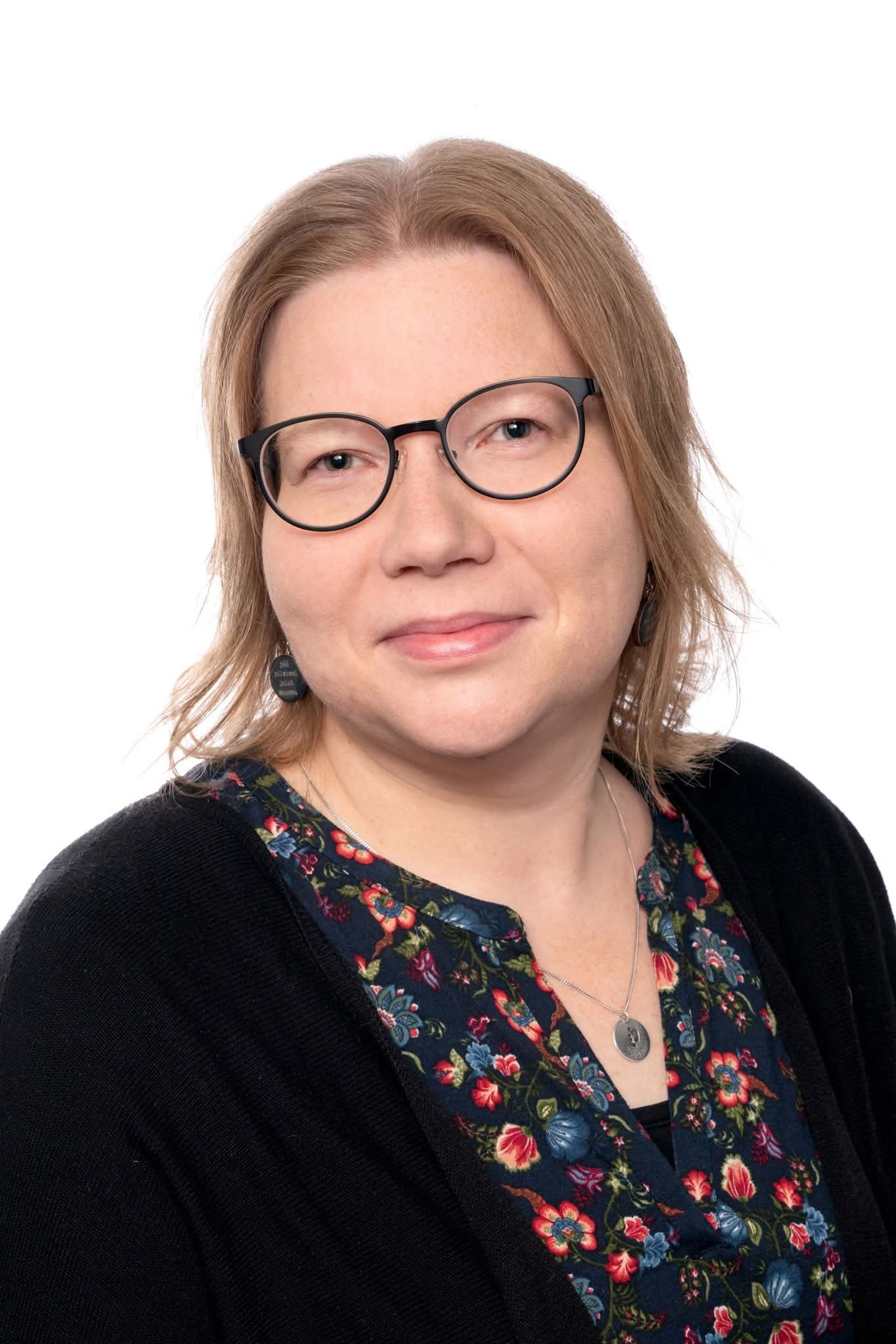 Kristiina Brisk‑Mosander katsoo kohti kameraa, päällään kuviollinen pusero, musta neuletakki ja silmälasit.