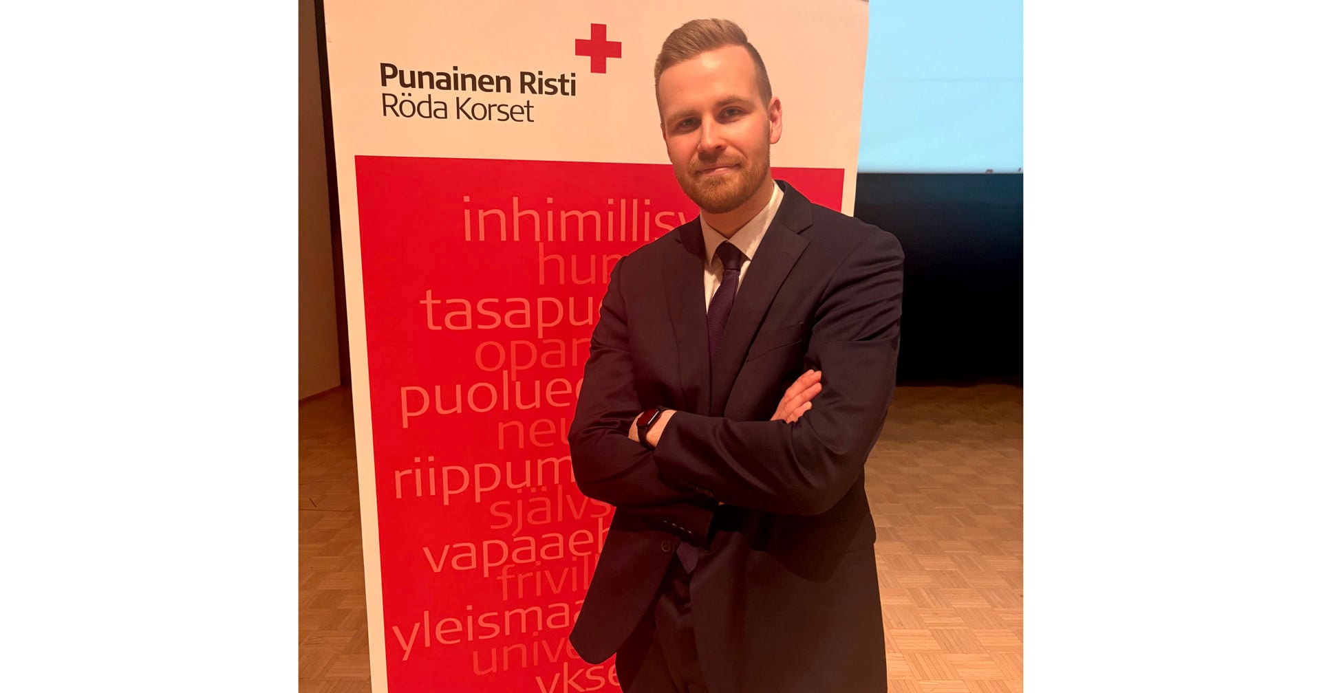 Mikko Sipilä seisoo kädet ristissä Punaisen Ristin banderollin edessä, päällään tumma puku ja solmio.