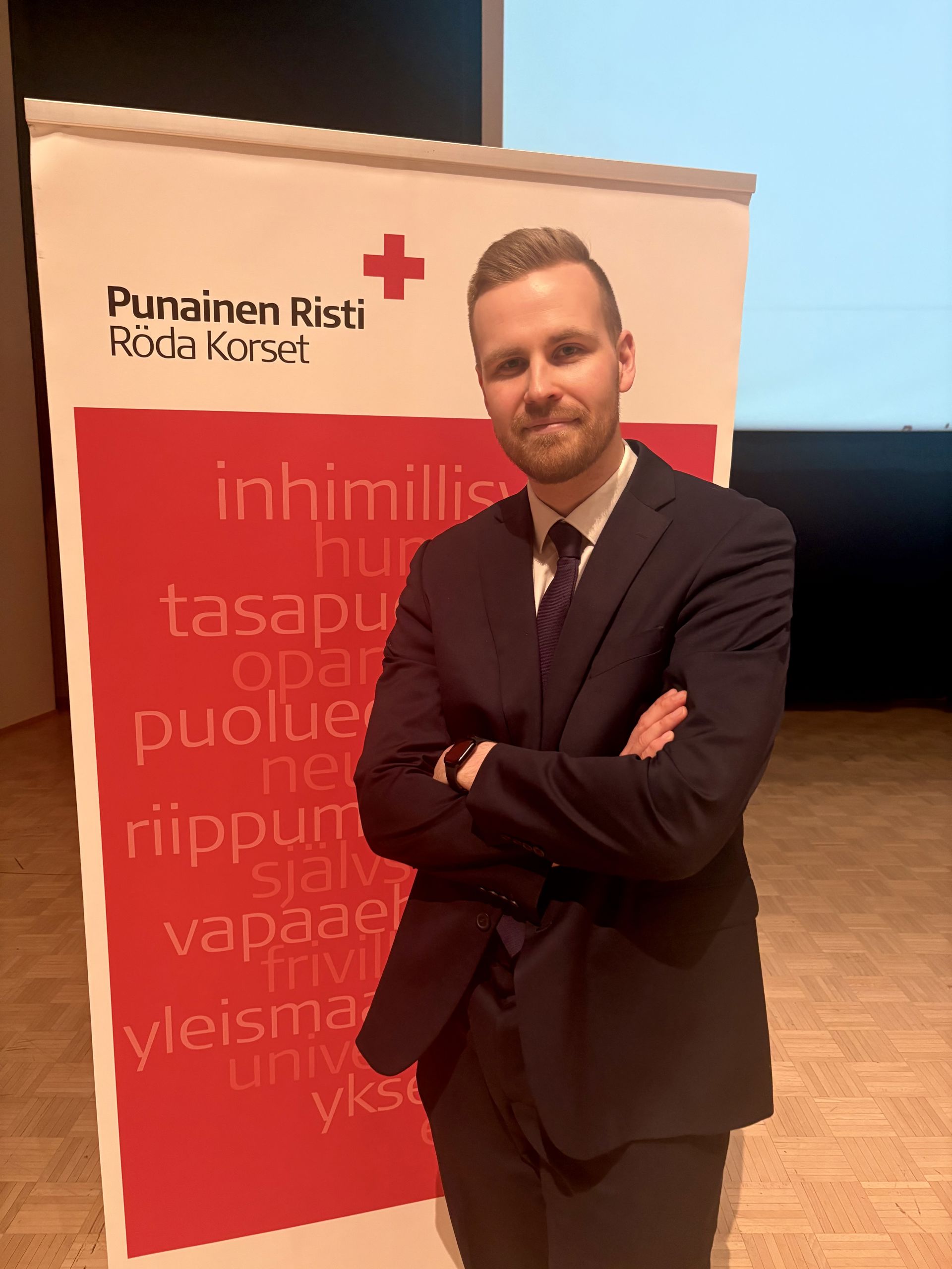 Mikko Sipilä seisoo kädet ristissä Punaisen Ristin banderollin edessä, päällään tumma puku ja solmio.