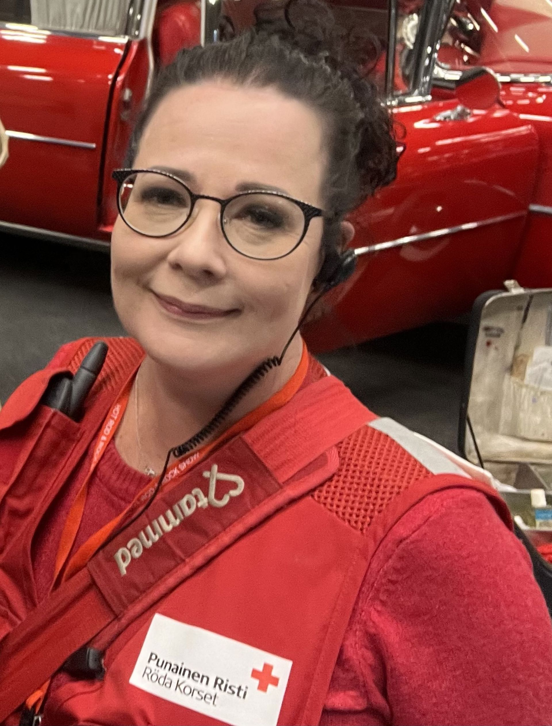 Niina Hokkanen katsoo kohti kameraa, päällään Punaisen Ristin liivit ja silmälasit. Taustalla on punainen ajoneuvo.