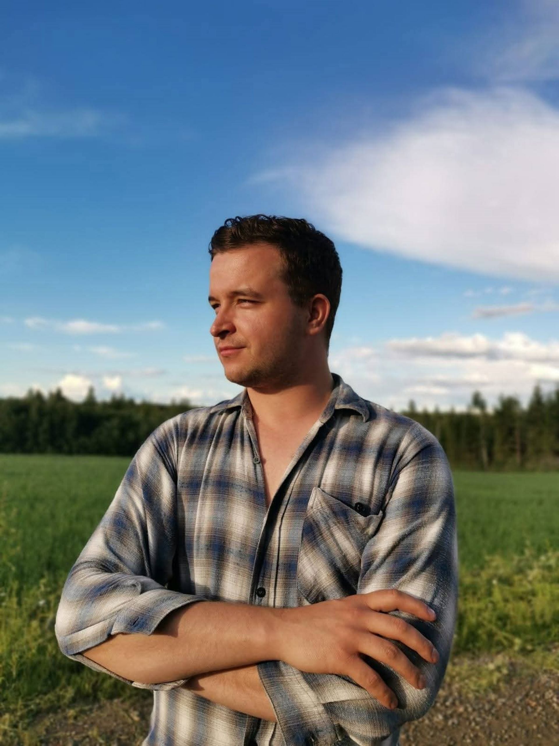 Osmo Tolonen seisoo kädet ristissä pellon laidalla, päällään ruutupaita. Taustalla näkyy vihreä pelto, metsänreuna ja sininen taivas.