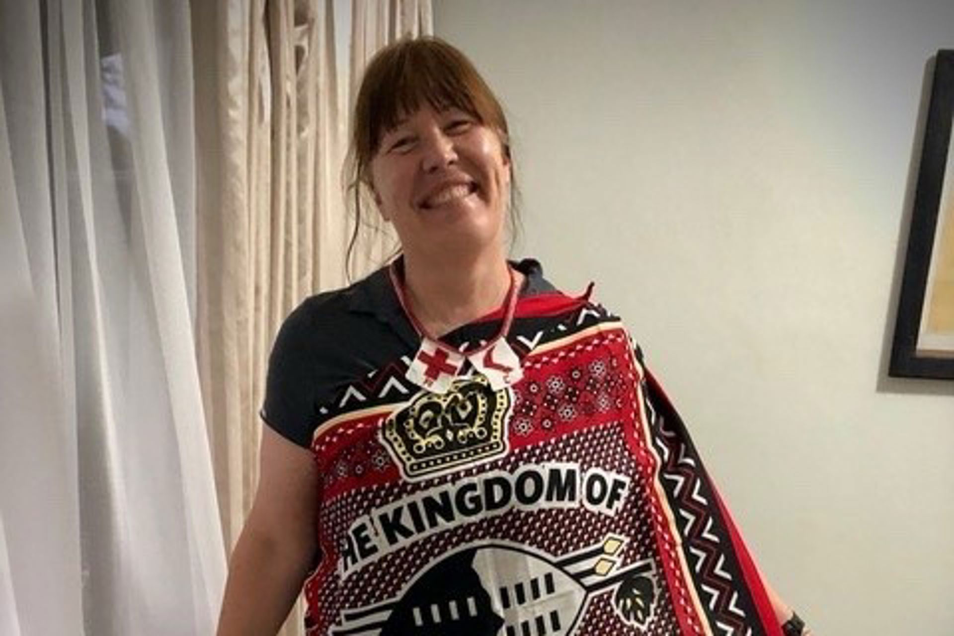 Päivi Hurttia seisoo sisätilassa vaalean verhon edessä, päällään tumma paita ja värikäs, punasävyinen kangas, jossa on kuvioita ja tekstiä, aseteltuna olkapäiden yli.