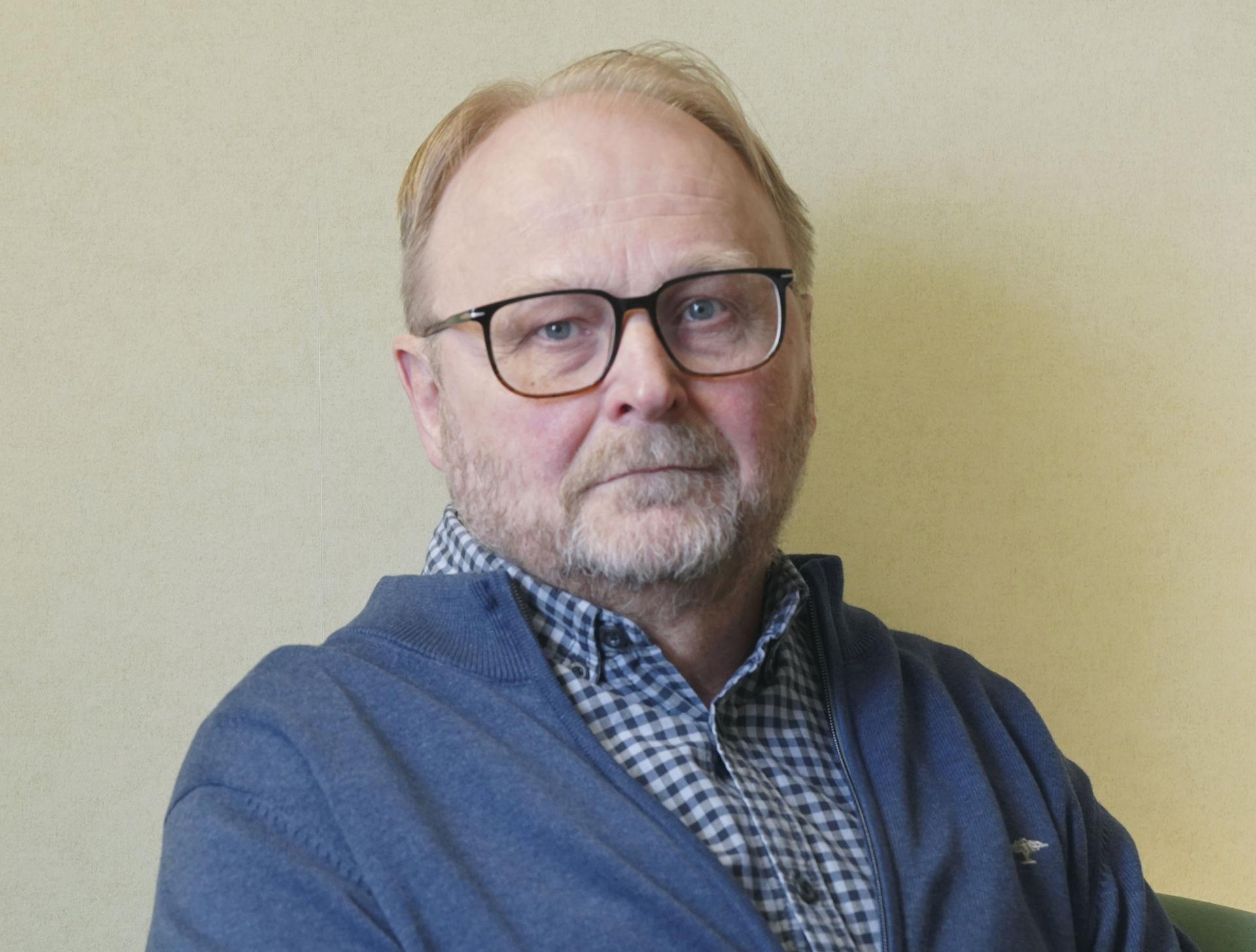Veli‑Matti Ahtiainen katsoo kohti kameraa, päällään ruudullinen kauluspaita, sininen neuletakki ja silmälasit.