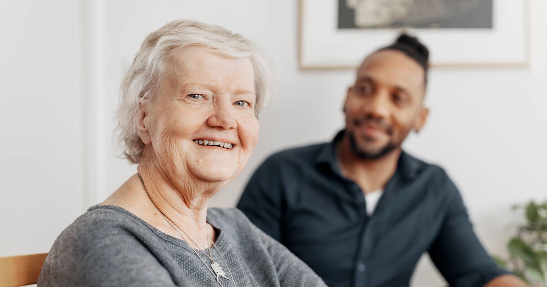 Vanhempi henkilö katsoo kameraan. Taustalla häntä hymyillen katsova henkilö.