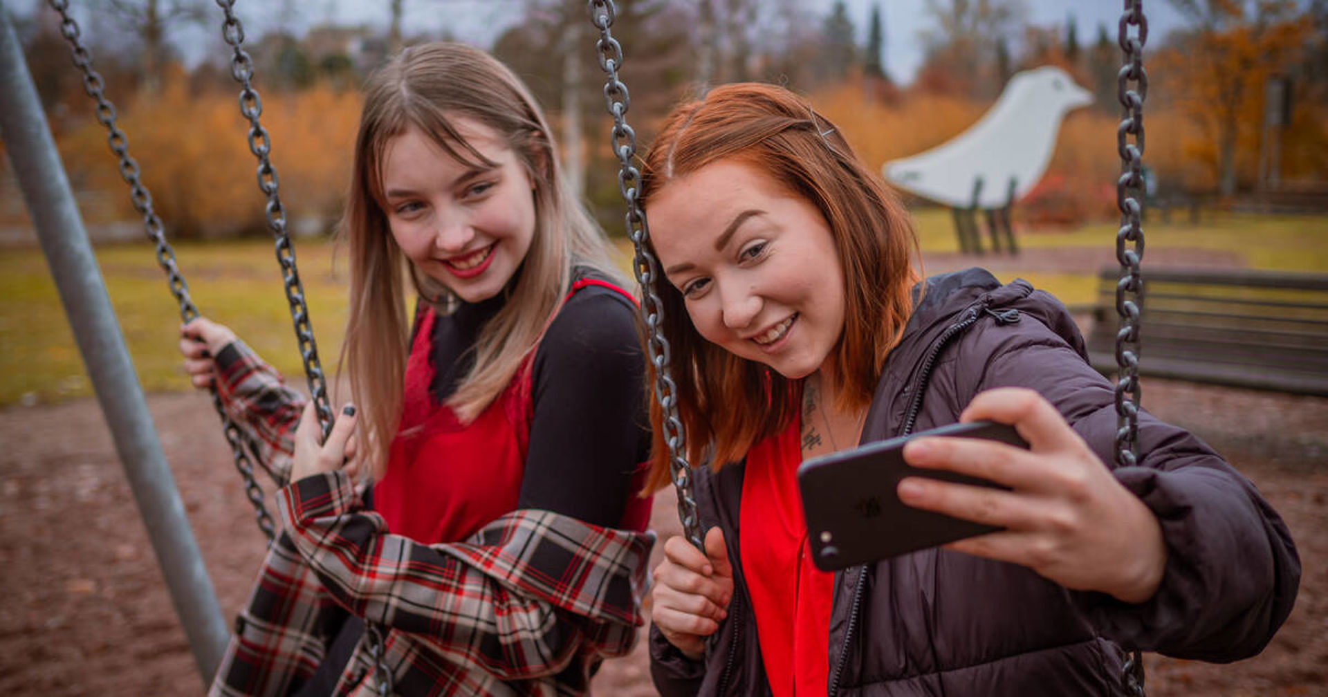 Kaksi nuorta ottaa kännykällä kuvaa keinuissa.