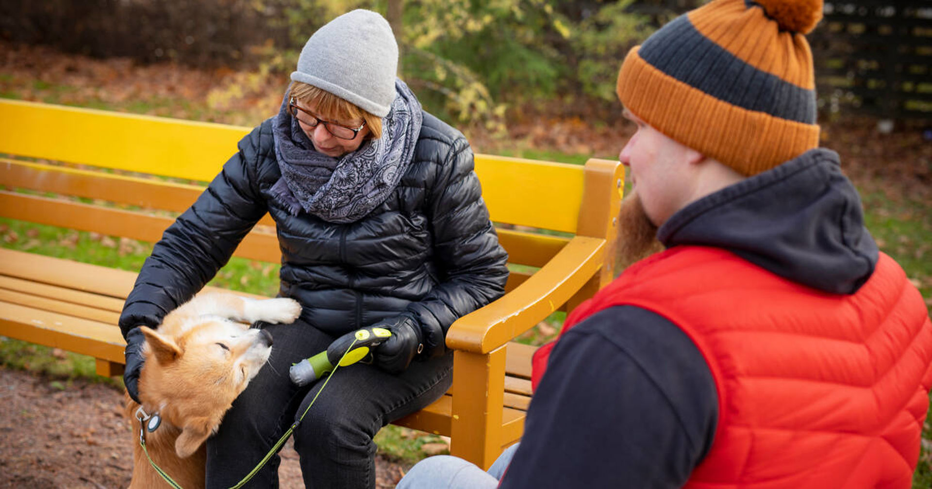 Kaksi henkilöä istuu ulkona. Toinen istuu penkillä ja toinen pyörätuolissa. Heillä on myös koira.